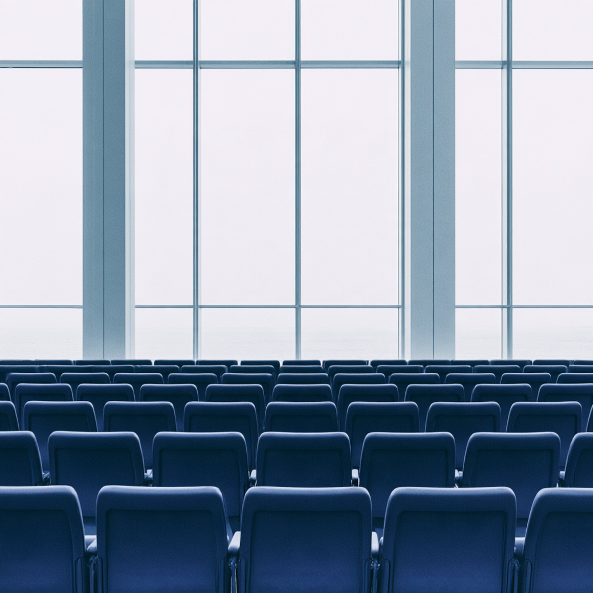 An empty lecture hall, rows of blue seats facing tall windows in cold light